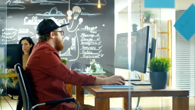 In-the-Creative-Agency-Videographer-in-a-Cap-Works-on-Personal-Computer-with-Two-Monitors,-He's-Doing-Video-and-Audio-Editing.-Stylish-Coworkers-at-Their-Workstations-and-Chalkboard-Wall-Visible.