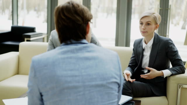 Panning-shot-of-businesswoman-talking-and-duscussing-with-male-business-colleagues-sitting-on-couch-in-modern-office-hall