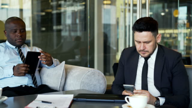 Dos-empresarios-multiétnicos-en-ropa-formal-sentado-y-usando-su-smartphone-y-tablet-en-café-vidrioso-durante-la-hora-del-almuerzo