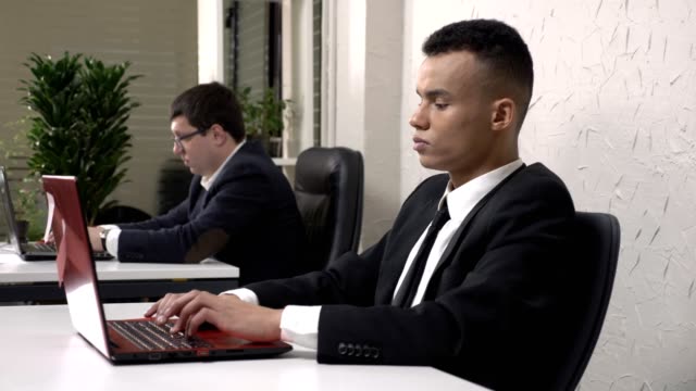 A-young-successful-African-male-businessman-working-in-the-office-on-a-laptop,-tired,-holding-his-head,-a-headache-concept.-Caucasian-man-in-suit-in-the-background.-60-fps