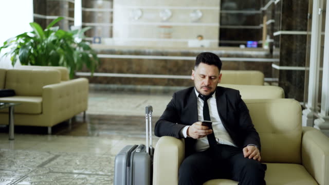 Pan-shot-of-concentrated-businessman-using-smartphone-sitting-on-armchair-inside-luxury-hotel-with-luggage-near-him