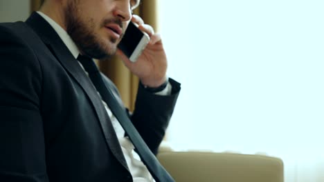 Close-up-tilt-up-of-confident-businessman-talking-mobile-phone-and-writing-notes-in-notepad-while-sits-on-armchair-in-hotel-room-or-office.-Travel,-business-and-people-concept