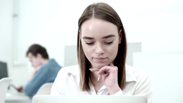 Primer-plano-de-hermosa-alumna-leyendo-en-la-pantalla-del-ordenador-portátil-y-sonriente,-su-estudiante-del-compañero-en-teléfono-de-fondo