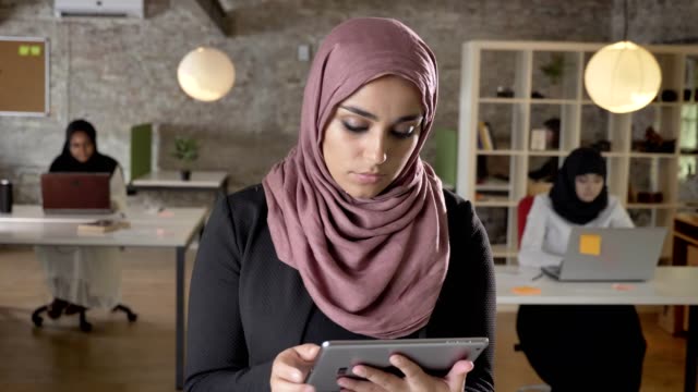 Portrait-of-young-muslim-women-in-hijab-standing-with-tablet,-looking-in-camera,-smiling,-business-womens-typing-on-laptop-in-office