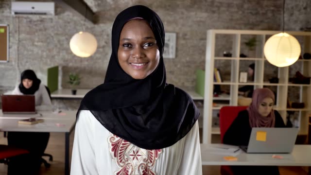 Junge-schwarze-Muslimas-Hijab-stehend-und-in-der-Kamera-suchen,-Lächeln,-drei-Frauen,-die-Arbeiten-am-Laptop-im-modernen-Büro