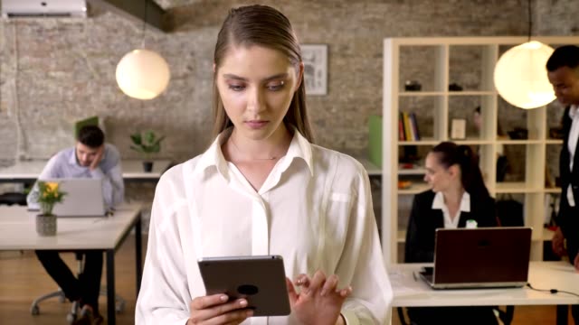 Young-concentrated-businesswoman-is-tapping-on-tablet-in-office,-watching-at-camera,-her-colleagues-are-networking-with-technologies,-working-concept,-business-concept,-communication-concept
