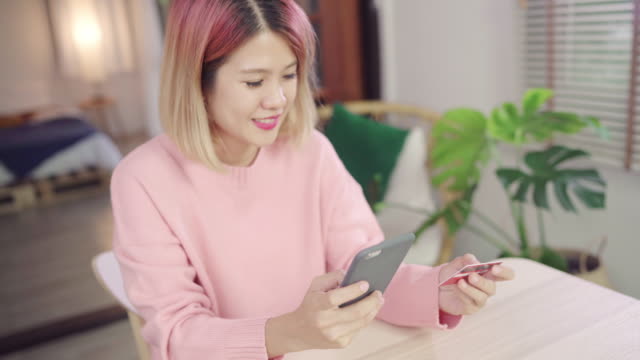 Beautiful-Asian-woman-using-smartphone-buying-online-shopping-by-credit-card-while-wear-sweater-sitting-on-desk-in-living-room-at-home.-Lifestyle-woman-at-home-concept.