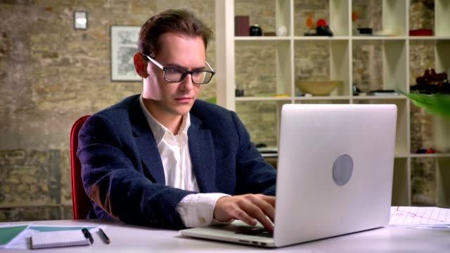 Concentrated-caucasian-male-is-sitting-and-looking-at-his-working-laptop-and-then-at-camera-relaxed-on-brick-background