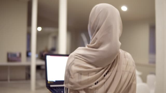 Joven-mujer-árabe-llevar-un-hijab-va-por-el-pasillo-mientras-mantiene-su-portátil-negro-en-las-manos.-Empleado,-lugar,-sala-de-conferencias,-pasillo.-Vista-de-la-parte-trasera,-fondo-desenfocado