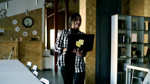 Caucasian-positive-woman-working-in-public-workplace-with-brick-wall-interior.-Beautiful-young-brunette-in-plaid-shirt-is-working-in-modern-workspace,-walking-by-room-with-laptop.-Smiling,-positive-business-woman-greets-colleagues