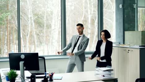 Pretty-businesswoman-is-teaching-her-male-coworker-to-dance-listening-to-music-and-moving-in-office-enjoying-break-from-work.-People-are-wearing-suits.