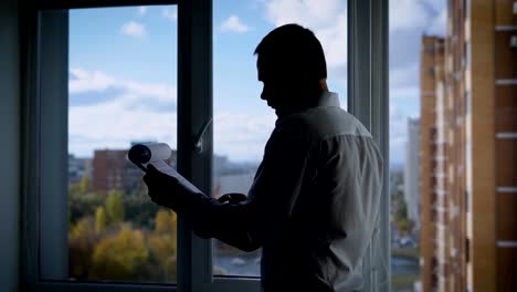 concentrated-businessman-is-reading-a-contract-on-a-paper-sheets,-standing-in-front-of-window-in-his-office-in-autumn-day