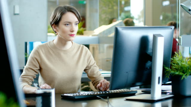 Hermosa-mujer-caucásica-joven-se-sienta-en-su-escritorio-y-trabaja-en-una-computadora-Personal.-En-el-fondo-oficina-ocupada-con-colegas-de-trabajo.