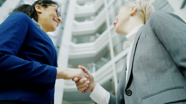 Low-angle-shot-of-businesswoman-shaking-hands-and-talking-with-female-business-colleague-in-suit-in-hall-of-modern-office-building