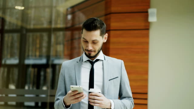 Stedicam-shot-of-cheerful-businessman-using-smartphone-and-walking-in-modern-office-hall