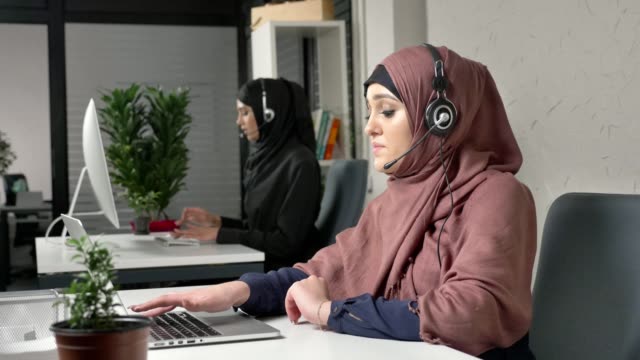 Una-joven-hermosa-en-un-hiyab-rosa-es-hablar-de-los-auriculares,-contestar-llamadas-en-call-center.-Mujeres-árabes-en-la-oficina.-60-fps