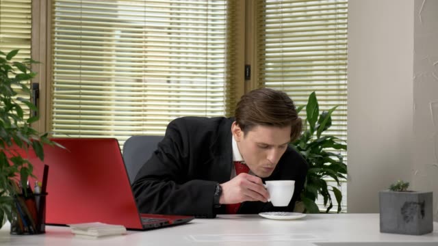young-guy-in-a-suit-is-sitting-in-the-office,-working-on-a-laptop,-not-drinking-tasty-coffee,-making-funny-faces.-Work-in-office.-60-fps