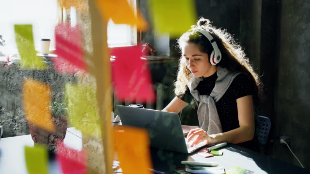 Attractive-female-student-is-listening-to-music-with-headphones-dancing-and-singing-while-working-with-laptop-computer.-Glassboard-with-bright-colored-stickers-in-foreground.