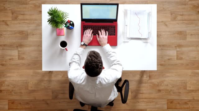 Hombre-joven-escribiendo-en-la-computadora-portátil-y-busca-en-la-pantalla,-topshot,-sentado-detrás-de-la-mesa-con-documentos-y-café