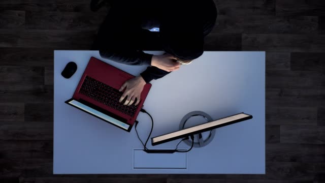 Young-man-in-black-hoodie-drinking-coffee-and-typing-on-laptop-during-night,-sitting-behind-white-desk,-top-shot