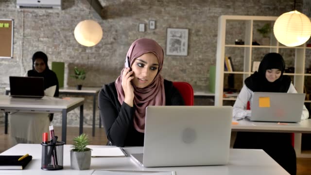 Three-young-muslim-womans-in-hijab-sitting-and-working-in-modern-office,-beautiful-muslim-woman-talking-on-phone,-smiling