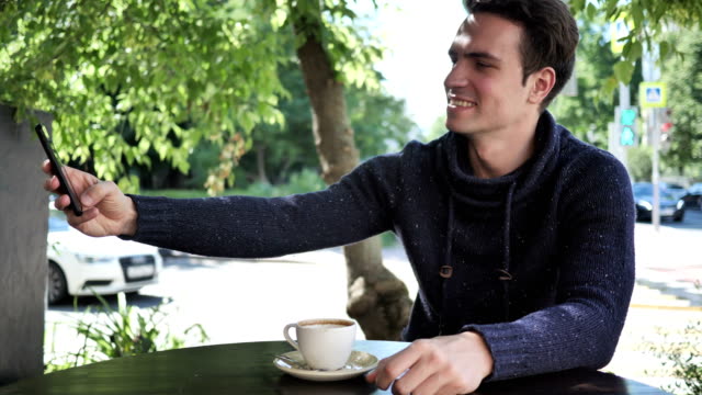 Young-Man-Taking-Selfie-on-Phone-while-Sitting-in-Cafe-Terrace