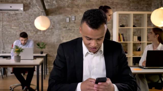Young-afro-american-businessman-is-typing-message-on-smartphone-in-office,-his-colleagues-are-networking-with-technologies,-working-concept,-business-concept,-communication-concept