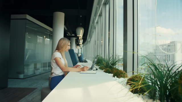 Empresaria-con-laptop-en-nueva-oficina-de-trabajo.-Vista-lateral-de-la-mujer-sentada-en-la-mesa-junto-a-ventana-en-oficina-moderna-y-usando-laptop-en-luz-del-día