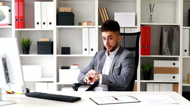 young-businessman-in-gray-jacket-sitting-at-table-in-white-office,-looking-at-clock-and-leaving-on-chair