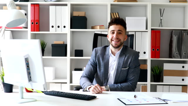 young-businessman-sitting-at-table-in-white-office,-smiling-and-showing-approving-hand-gesture