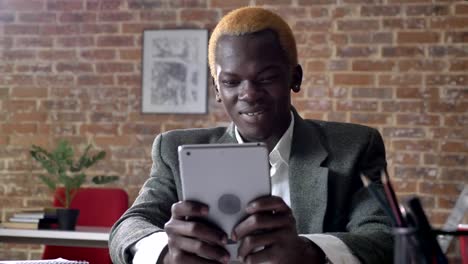 Young-african-american-blonde-businessman-browsing-on-tablet-and-smiling,-sitting-in-modern-office,-confident-and-successful