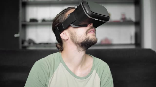 Young-bearded-hipster-man-using-his-VR-headset-display-for-virtual-reality-game-or-watching-the-360-video-while-sitting-on-sofa-at-home-in-the-living-room.-VR-Technology.