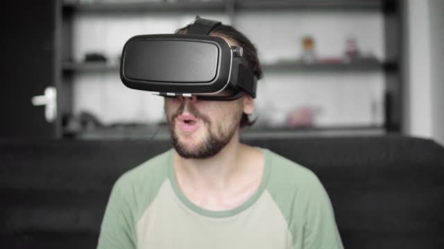 Young-bearded-hipster-man-using-his-VR-headset-display-for-watching-the-360-video-while-sitting-on-sofa-and-eating-cookies-at-home-in-the-living-room.-VR-Technology.