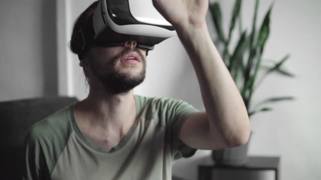 Young-bearded-hipster-man-using-his-VR-headset-display-for-virtual-reality-game-or-watching-the-360-video-and-trying-to-touch-to-something-he-see-while-sitting-on-sofa.-VR-Technology.