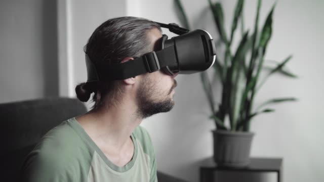 Joven-barbudo-a-hombre-hipster-usando-su-pantalla-del-auricular-VR-para-juego-de-realidad-virtual-o-ver-el-video-360-mientras-está-sentado-en-el-sofá-en-casa-en-la-sala-de-estar.-Tecnología-VR.