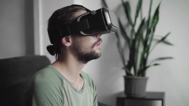 Young-bearded-hipster-man-using-his-VR-headset-display-for-virtual-reality-game-or-watching-the-360-video-and-trying-to-touch-to-something-he-see-while-sitting-on-sofa.-VR-Technology.