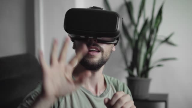 Young-bearded-hipster-man-using-his-VR-headset-display-for-virtual-reality-game-or-watching-the-360-video-and-trying-to-drive-away-what-he-sees-while-sitting-on-sofa.-VR-Technology.