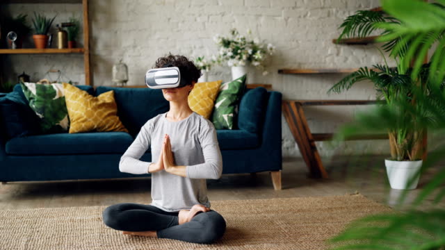 Slender-girl-in-virtual-reality-glasses-is-meditating-sitting-on-floor-at-home-in-lotus-position-and-enjoying-personal-practice.-Modern-technology-and-yoga-concept.