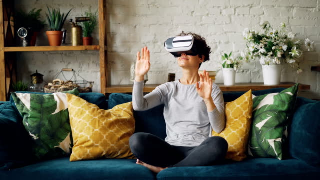 Zoom-in-of-smiling-young-lady-in-augmented-reality-glasses-moving-hands-and-turning-head-sitting-on-sofa-at-home-and-having-fun.-Entertainment-and-technology-concept.