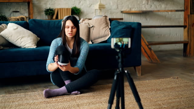 Attractive-lady-vlogger-is-using-smartphone-camera-to-record-video-about-virtual-reality-glasses.-Girl-is-holding-device,-showing-it-to-subscribers-and-talking.