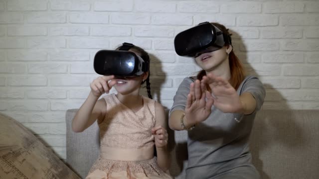 Two-young-girls-in-black-virtual-reality-glasses-sit-and-play-virtual-games.-They-wave-their-hands,-press-virtual-buttons-and-explore-the-surrounding-space.