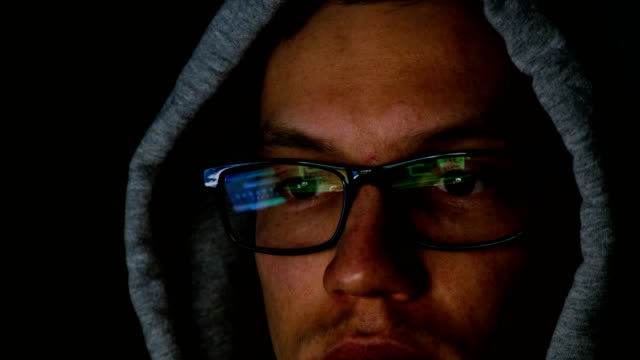 Hacker-in-glasses-and-a-hood-works-behind-a-computer-in-the-dark,-the-program-code-is-reflected-in-glasses-close-up,-4k