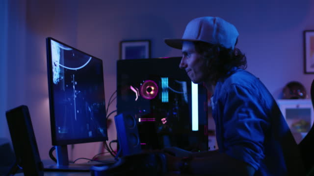 Excited-Gamer-Playing-and-Winning-in-First-Person-Shooter-Online-Video-Game-on-His-Personal-Computer.-Room-and-PC-have-Colorful-Neon-Led-Lights.-Young-Man-is-Wearing-a-Cap.-Cozy-Evening-at-Home.