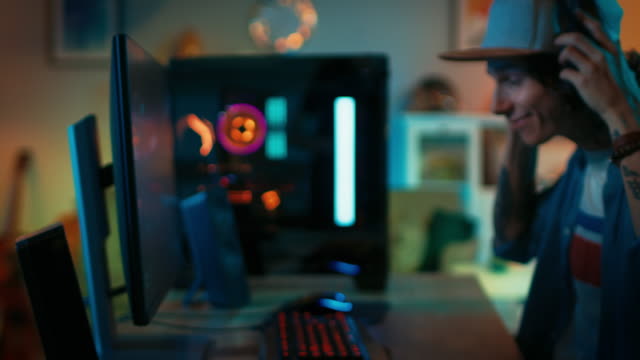 Gamer-Puts-His-Headset-with-a-Mic-On-and-Starts-Playing-Shooter-Online-Video-Game-on-His-Personal-Computer.-Room-and-PC-have-Colorful-Neon-Led-Lights.-Young-Man-is-Wearing-a-Cap.-Cozy-Evening-at-Home.