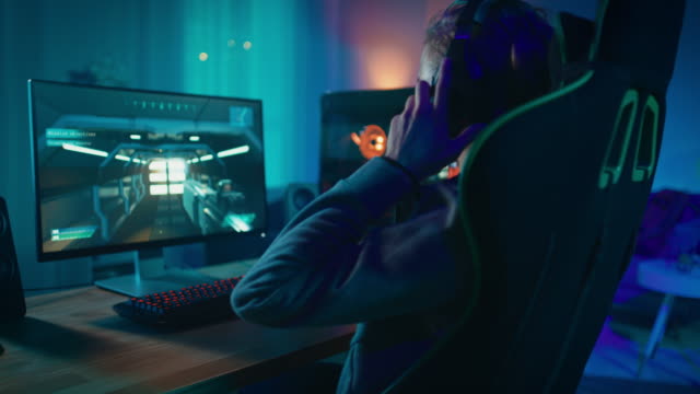 Spieler-setzt-seine-Kopfhörer-mit-Mikrofon-auf-und-startet-die-Wiedergabe-von-Online-Shooter-Videospiel-auf-seinem-persönlichen-Computer.-Zimmer-und-PC-haben-bunte-Neonlichter-geführt.-Gemütlichen-Sie-Abend-zu-Hause.