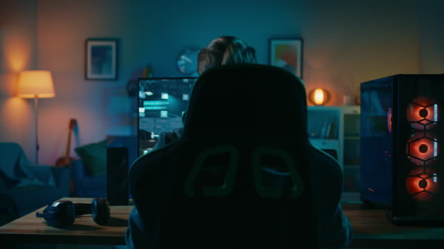 Back-Shot-of-a-Gamer-Playing-and-Winning-in-First-Person-Shooter-Online-Video-Game-on-His-Powerful-Personal-Computer.-Room-and-PC-have-Colorful-Neon-Led-Lights.-Cozy-Evening-at-Home.
