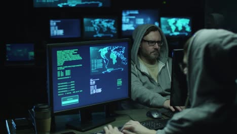 Two-hackers-in-hoods-work-on-a-computers-with-maps-and-data-on-display-screens-in-a-dark-office-room.