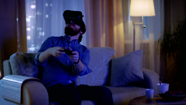 Man-Sitting-on-a-Couch-in-His-Living-Room--Plays-Video-Games-on-His-Console-While-Wearing-Virtual-Reality-Headset.