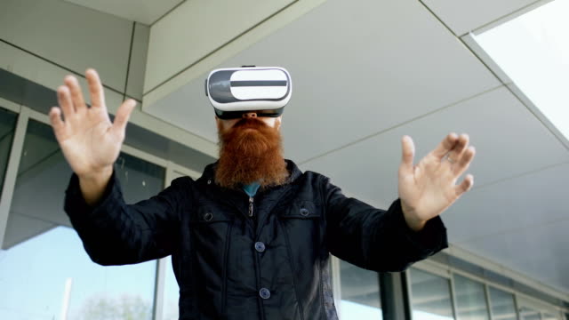 Young-bearded-man-using-virtual-reality-headset-for-360-VR-experience-while-walking-down-city-street-outdoors