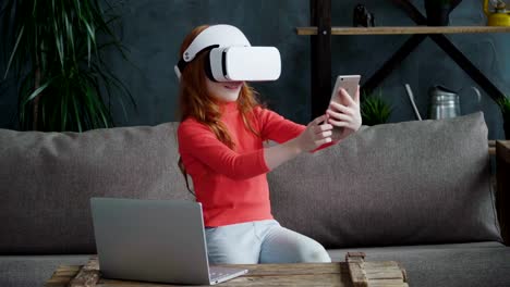 Little-redhead-girl-is-sitting-on-the-sofa-in-vr-headset-and-manipulate-virtual-reality-from-her-smartphone